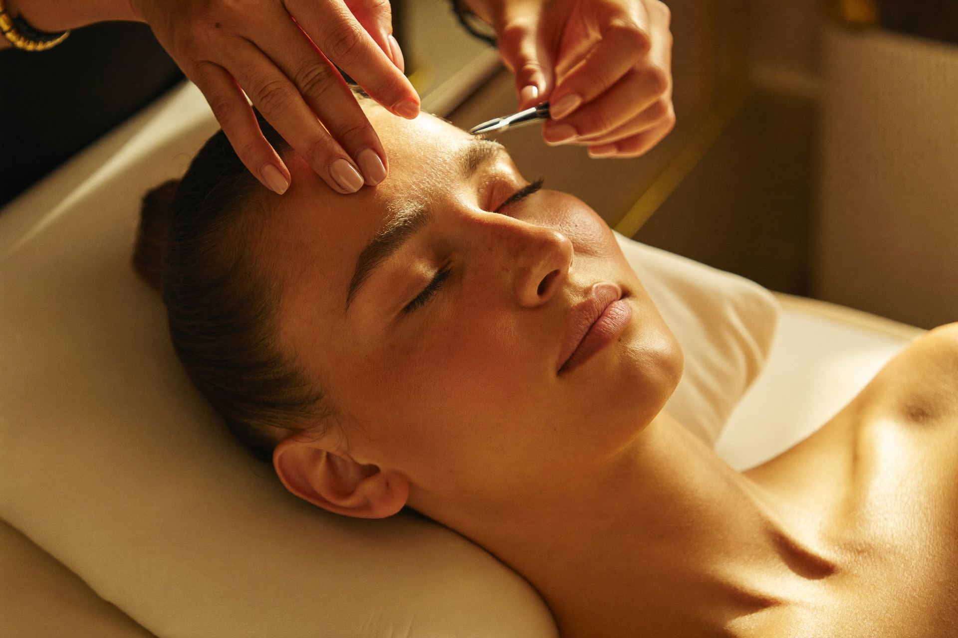Esthetician carefully shaping a client's eyebrow with tweezers in soft golden light
